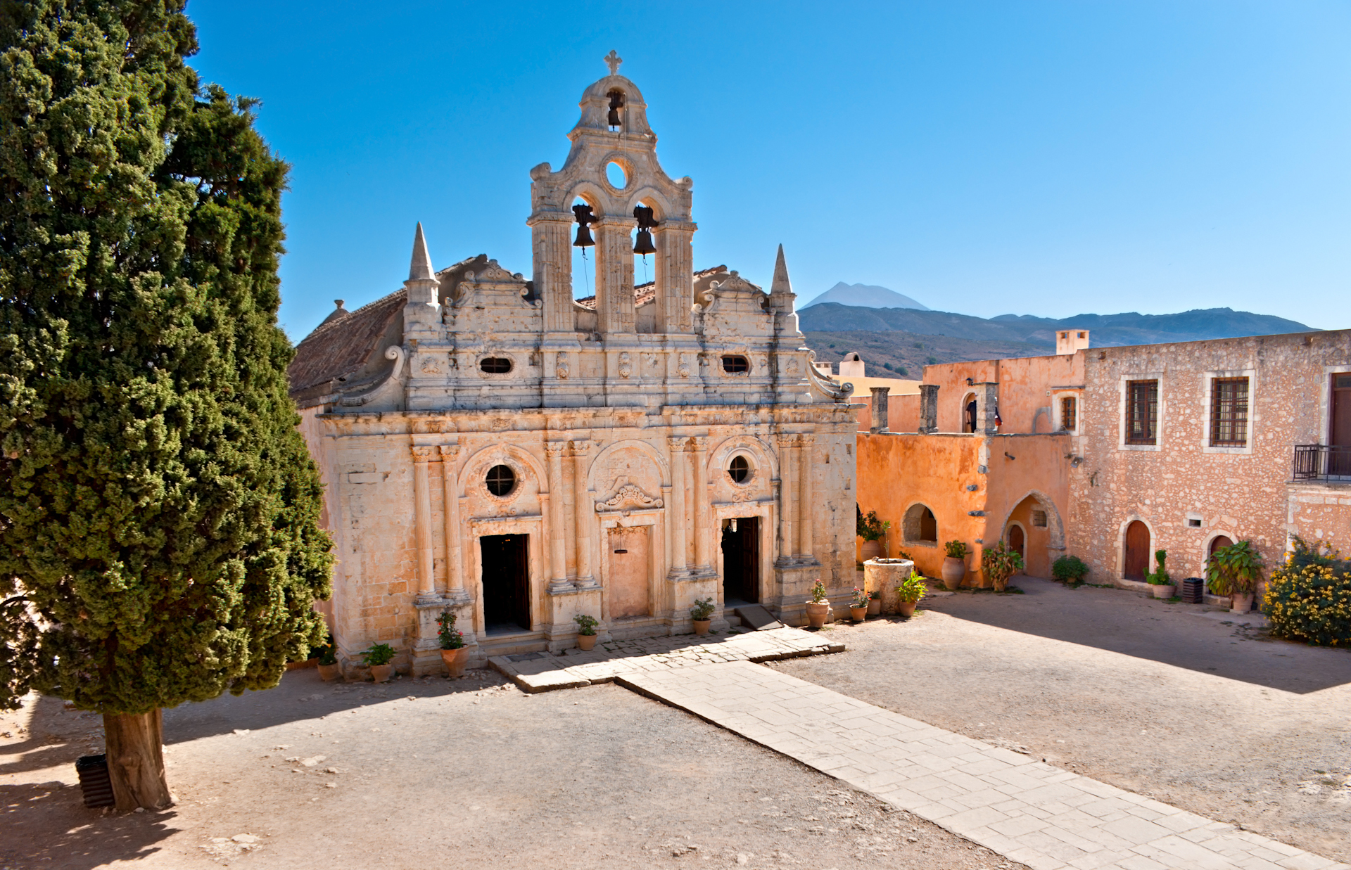 The historic Arkadi Monastery in Crete, showing the iconic Venetian-style church facade and surrounding courtyard under clear blue sky
