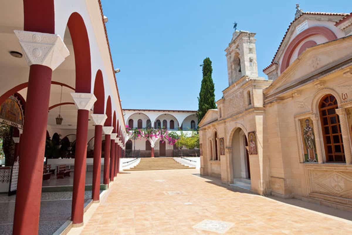The courtyard of Panagia Kalyviani Monastery in Crete, with red colonnades, stone church facade, bell tower and bright summer sky